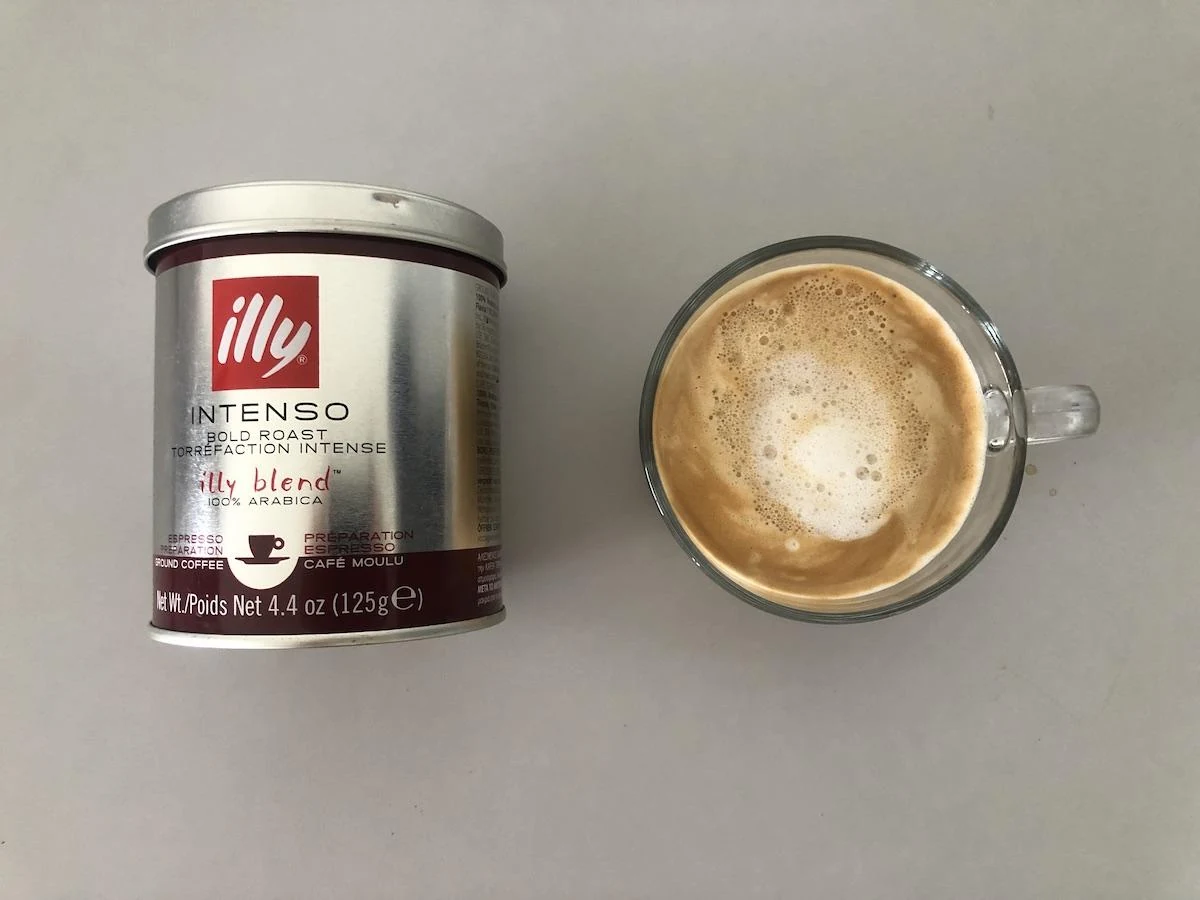 Illy Intenso ground espresso tin beside a glass cup of coffee with milk