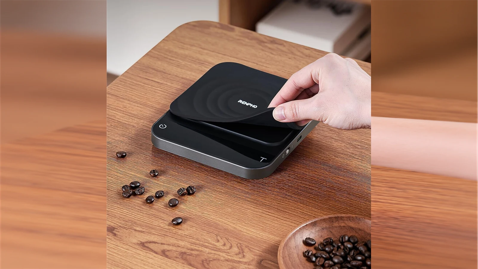 A hand lifting the silicone pad from a RENPHO Calibarista coffee scale on a wooden counter