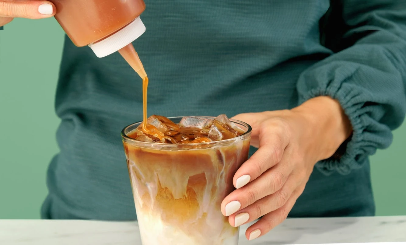 A hand adding a caramel drizzle over an iced milk coffee