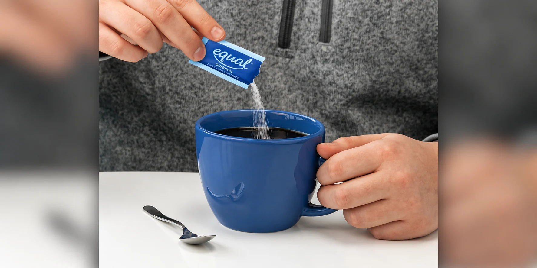 A hand pouring an Equal Original packet into a blue coffee mug