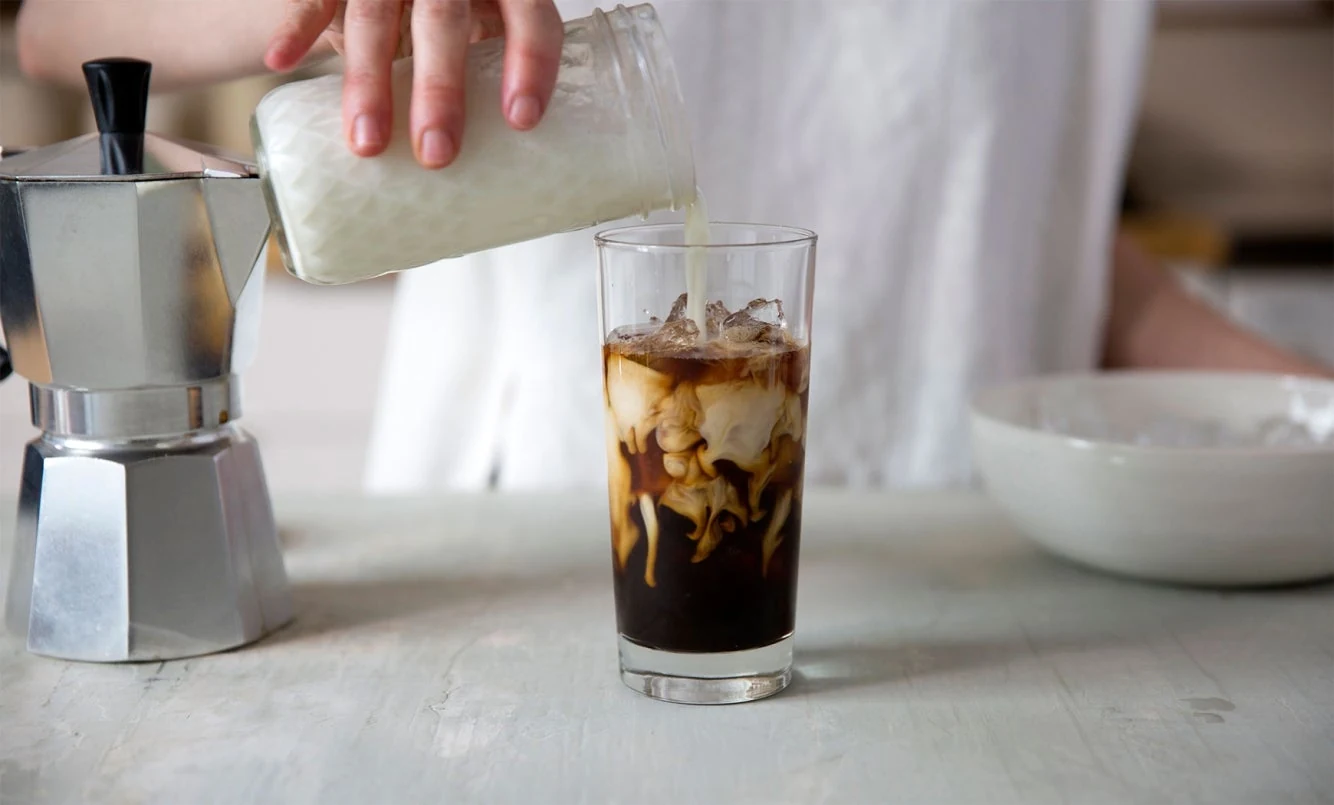 A hand pouring milk into a glass of iced coffee beside a moka pot