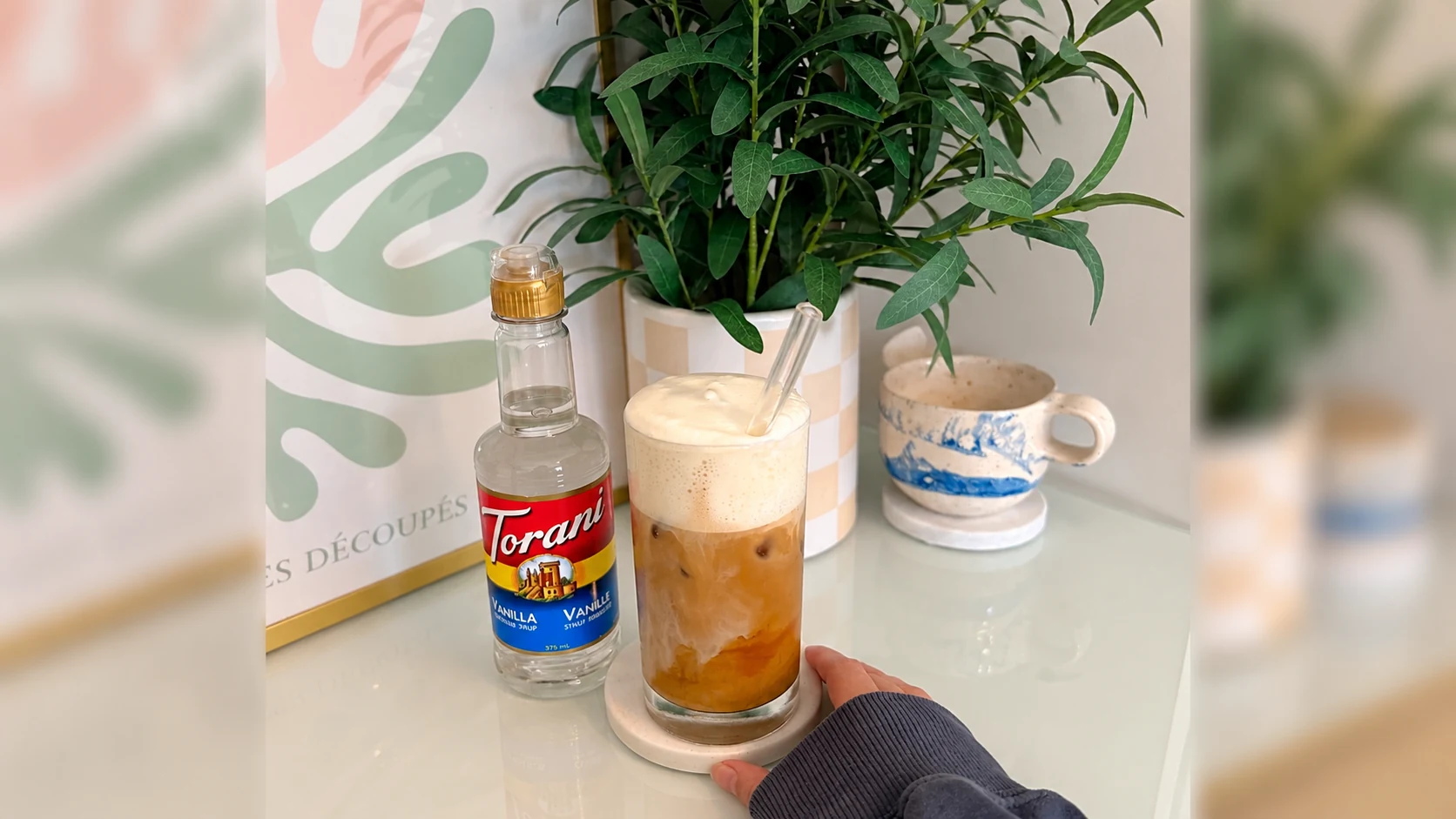 A hand holding an iced vanilla latte beside a smaller Torani Vanilla bottle
