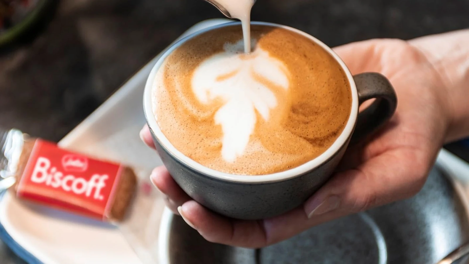 A wrapped Lotus Biscoff biscuit beside a latte while milk is being poured