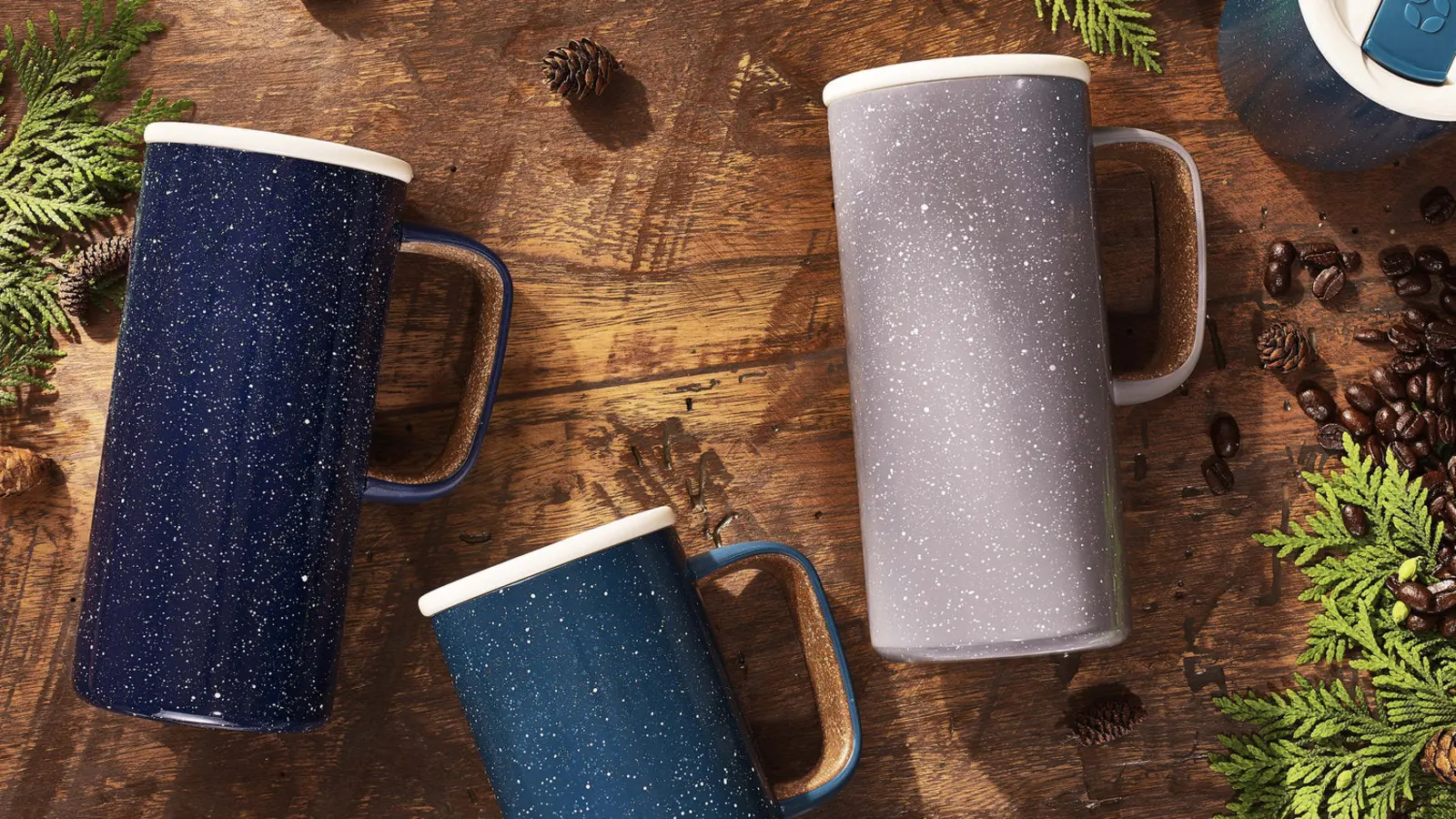 Three Ello Campy 18 oz travel mugs arranged on a wooden table with coffee beans