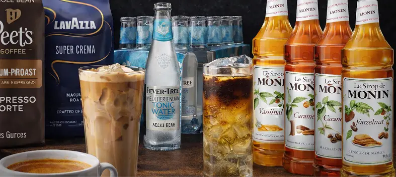 Espresso tonic and iced coffee drinks on a counter with tonic water bottles, flavored syrup bottles, and espresso coffee bags in the background.